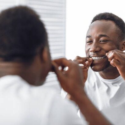 Toothy African Man Flossing And Cleaning Teeth Using Tooth Floss Standing Near Mirror In Modern Bathroom At Home In The Morning. Toothcare And Healthy Oral Hygiene Concept. Selective Focus